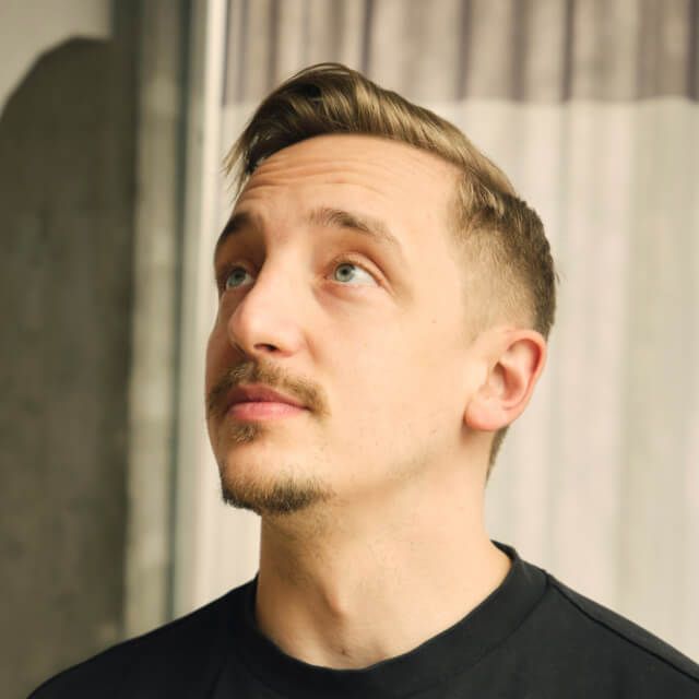 Person with short light brown hair, wearing a black shirt, standing indoors near a window