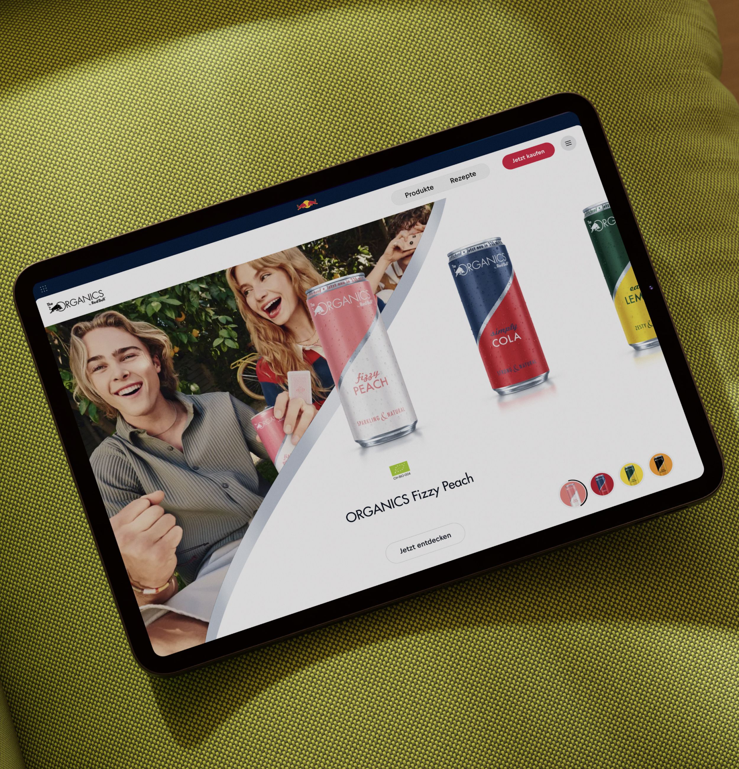 A tablet displaying a website with colorful cans of ORGANICS drinks and people holding beverages, placed on a green textured surface