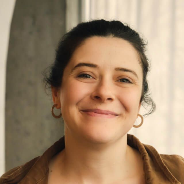 Person with dark curly hair wearing a brown jacket and hoop earrings, standing indoors