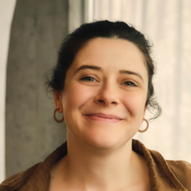 Person with dark curly hair wearing a brown jacket and hoop earrings, standing indoors