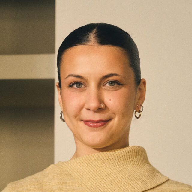 Person with dark hair tied back, wearing a beige turtleneck sweater and hoop earrings, standing indoors