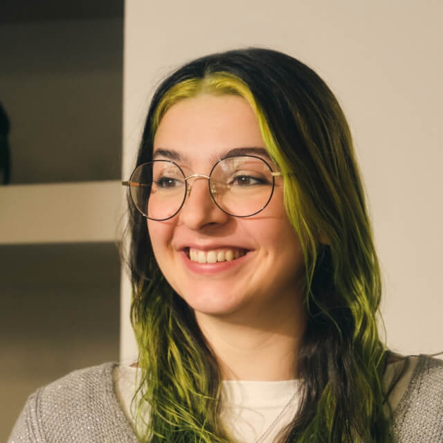 Person with long dark hair with green highlights, wearing glasses and a grey sweater, indoors