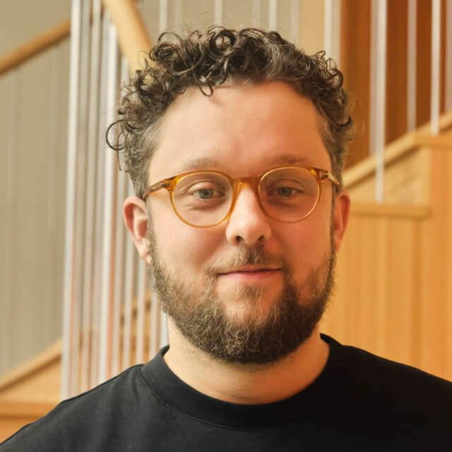 Person with short curly hair and beard, wearing a black shirt, standing in front of a wooden staircase