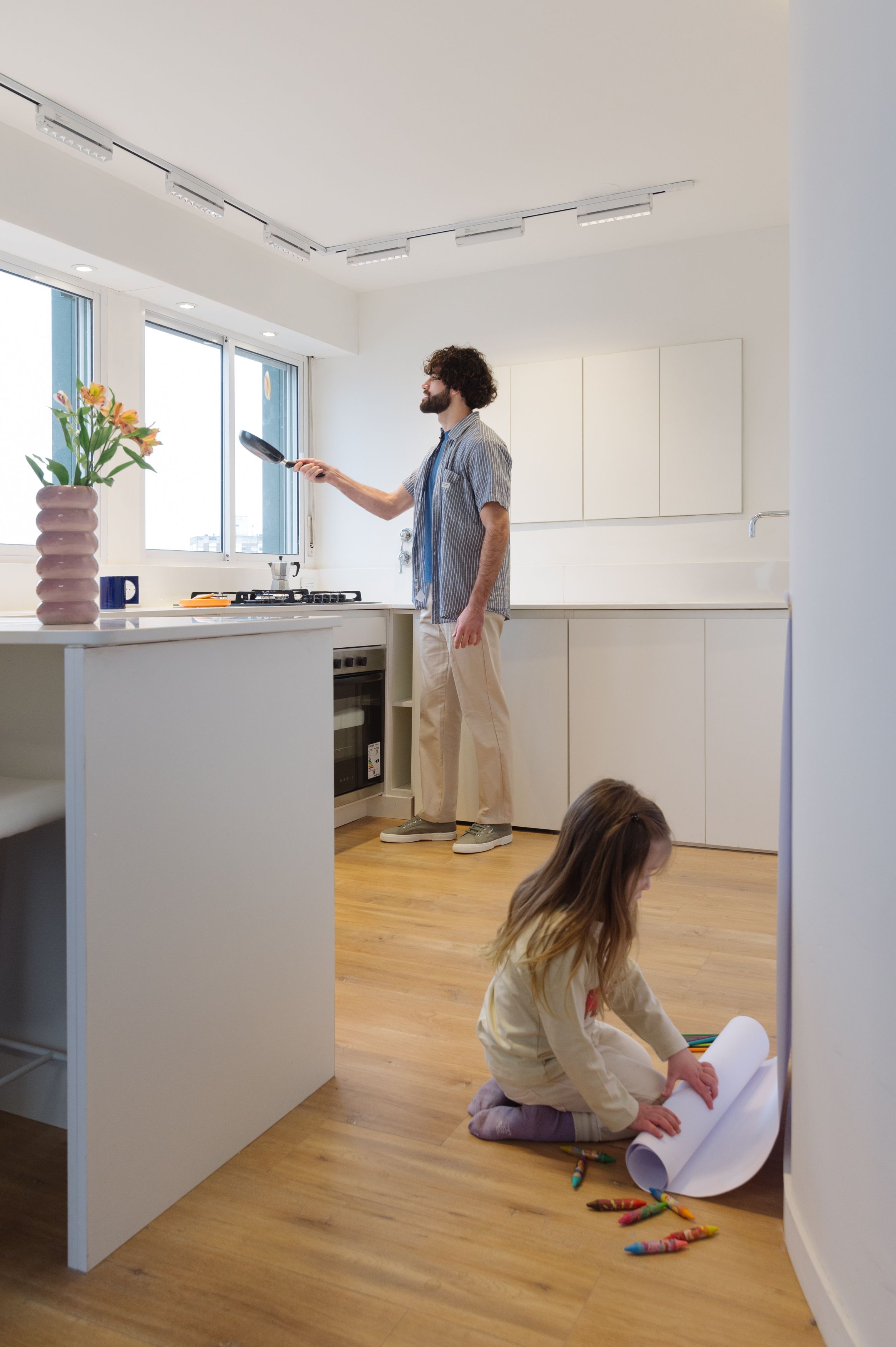 Cocina blanca de Y485 con un hombre cocinando y una niña dibujando en el piso de madera.