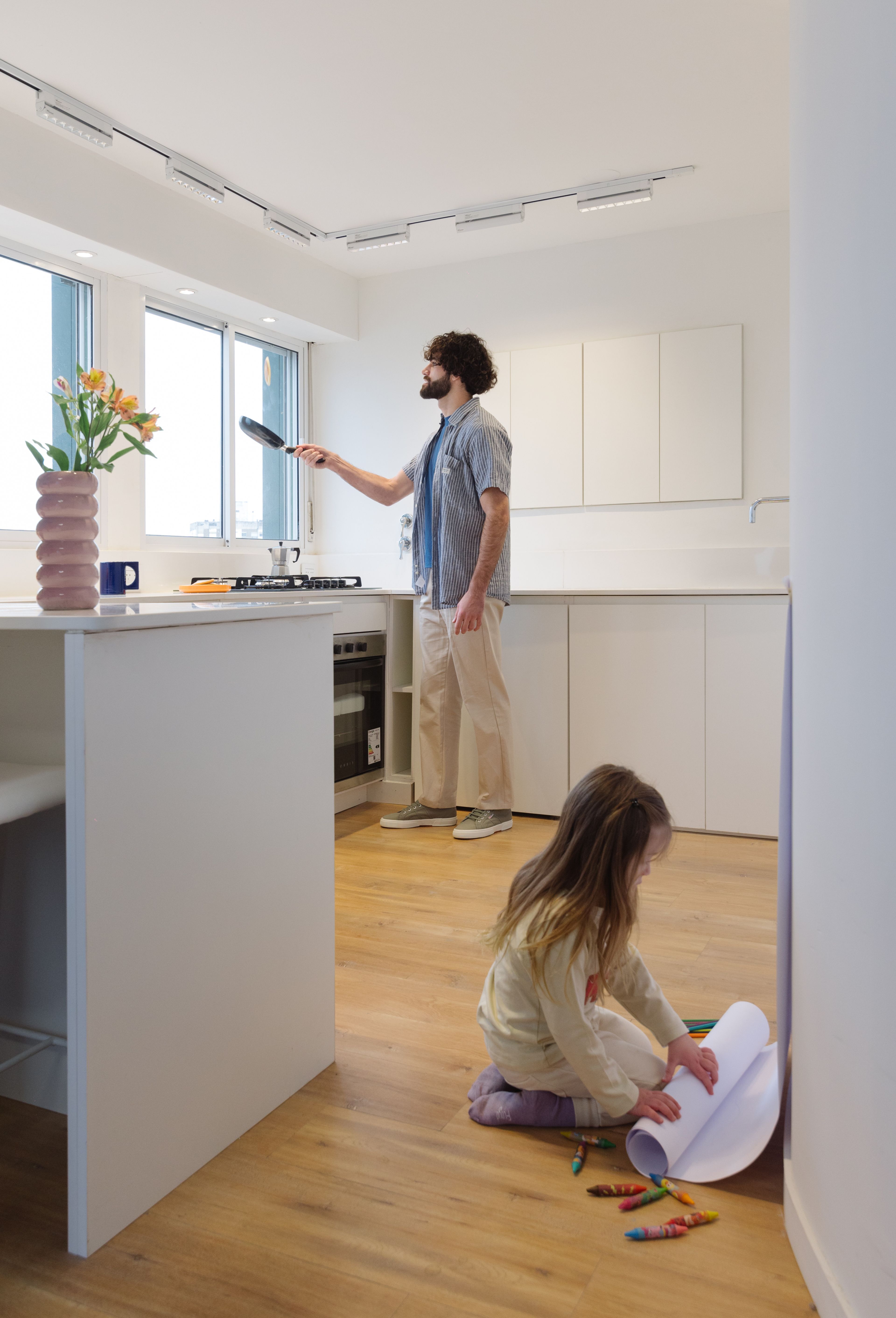 Cocina blanca de Y485 con un hombre cocinando y una niña dibujando en el piso de madera.