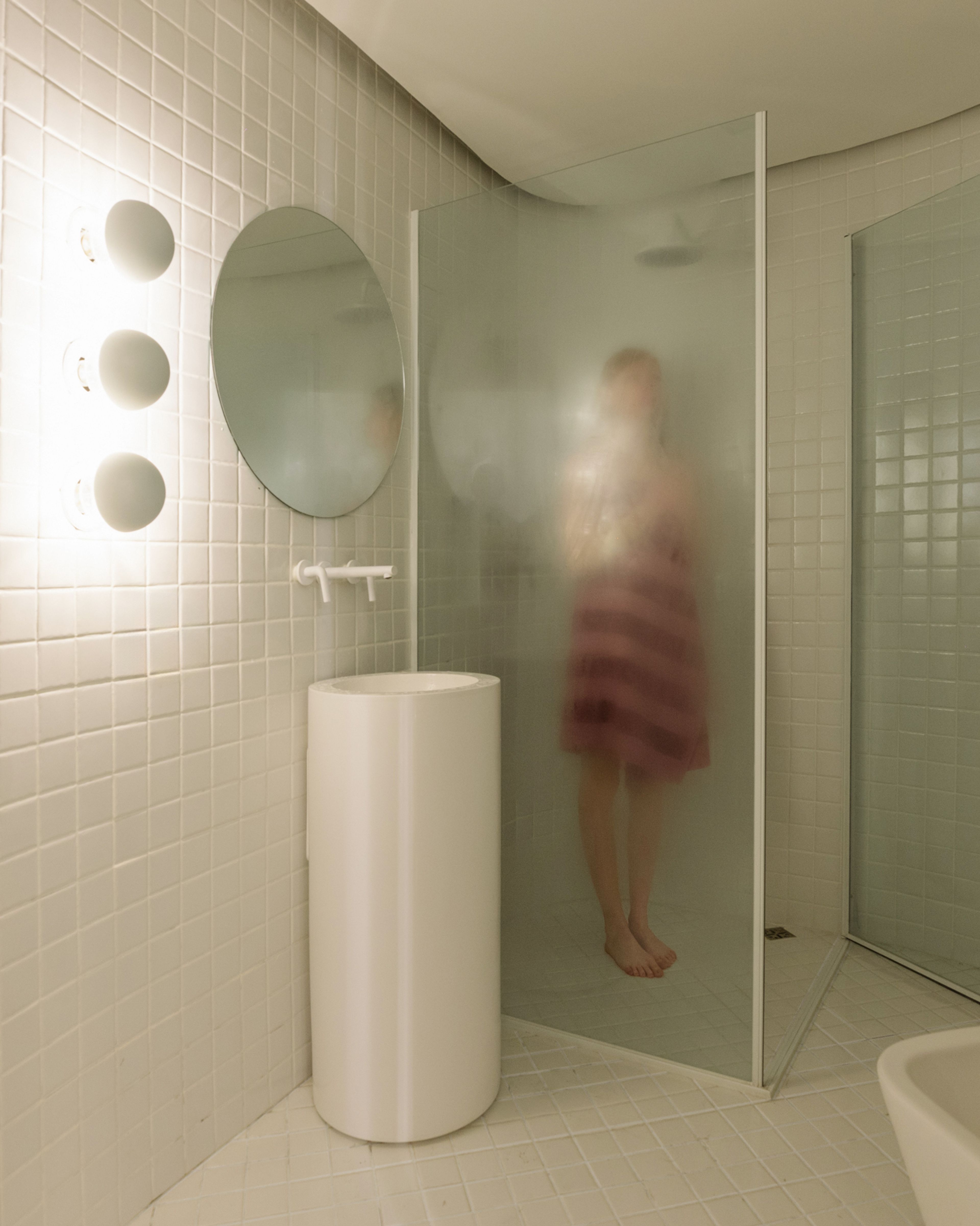 Baño de Y485 revestido en venecitas blancas, con lavabo cilíndrico, espejo redondo y ducha vidriada.