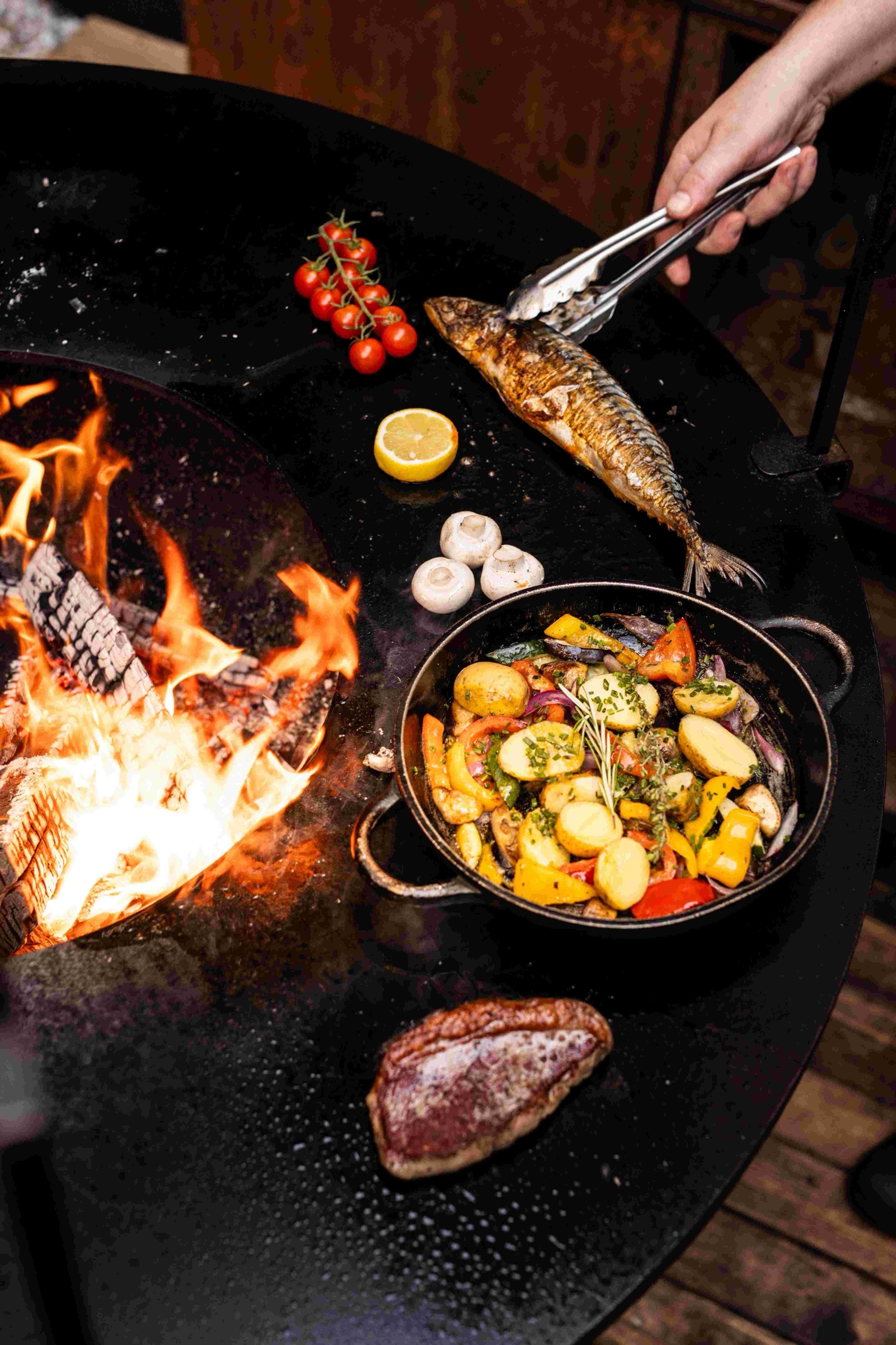 Ryba, steak a pánev se zeleninou na grilu nad otevřeným ohněm