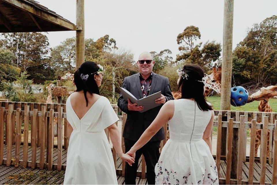 A wedding ceremony at Orana, with two brides and a celebrant on a wooden deck