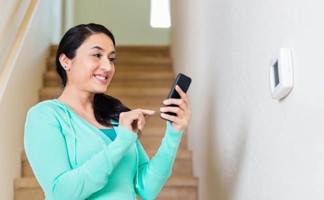 woman standing by thermostat using her phone to control it