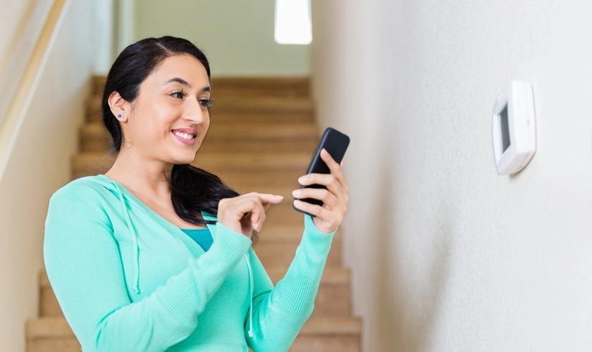 woman standing by thermostat using her phone to control it
