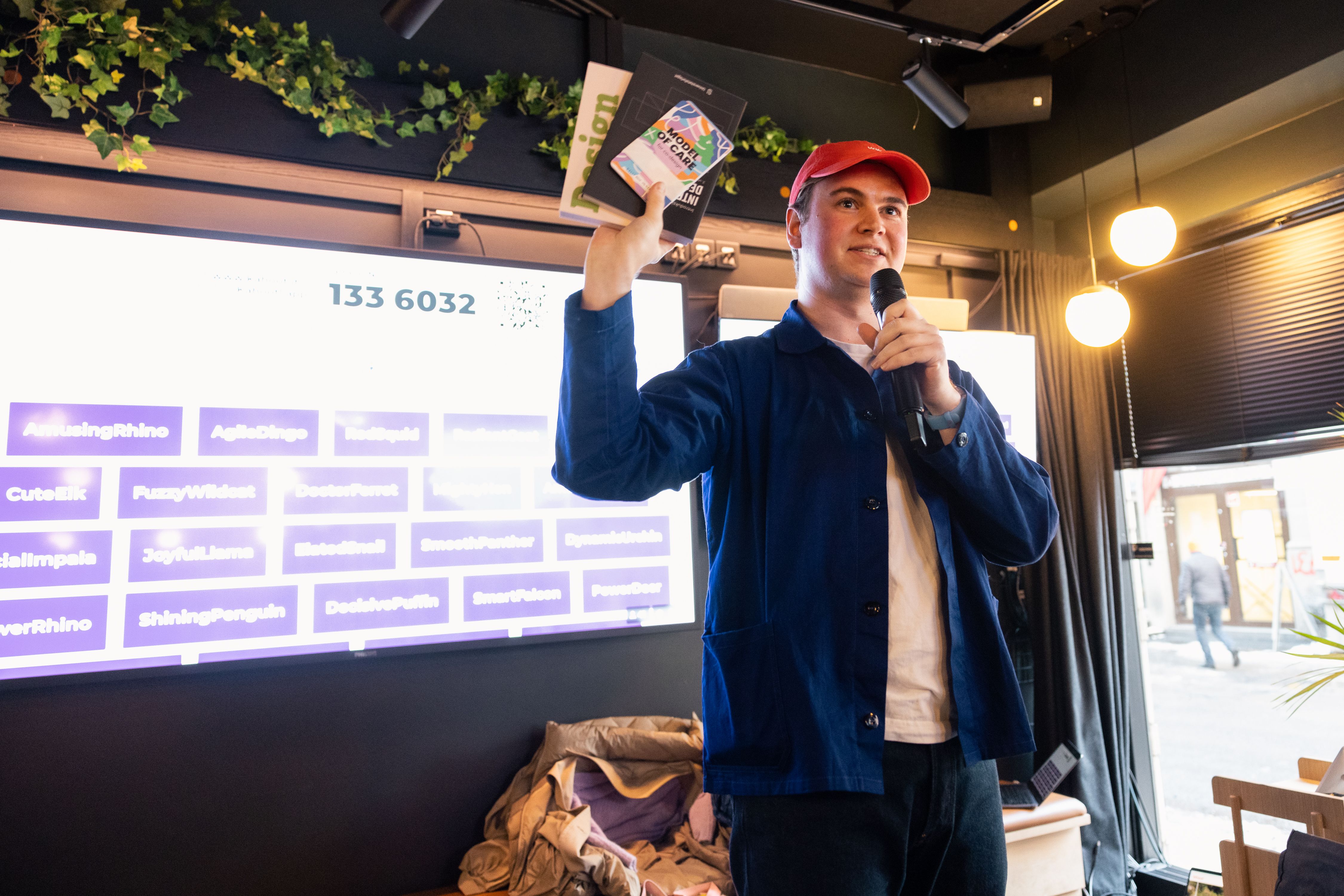 Jens Obel of IxDA Oslo stands onstage at MESH Youngstorget with two design books and stack of method cards in his right hand, while speaking into an handheld mic. Behind him, a large screen shows the names of the participants in the upcoming Kahoot