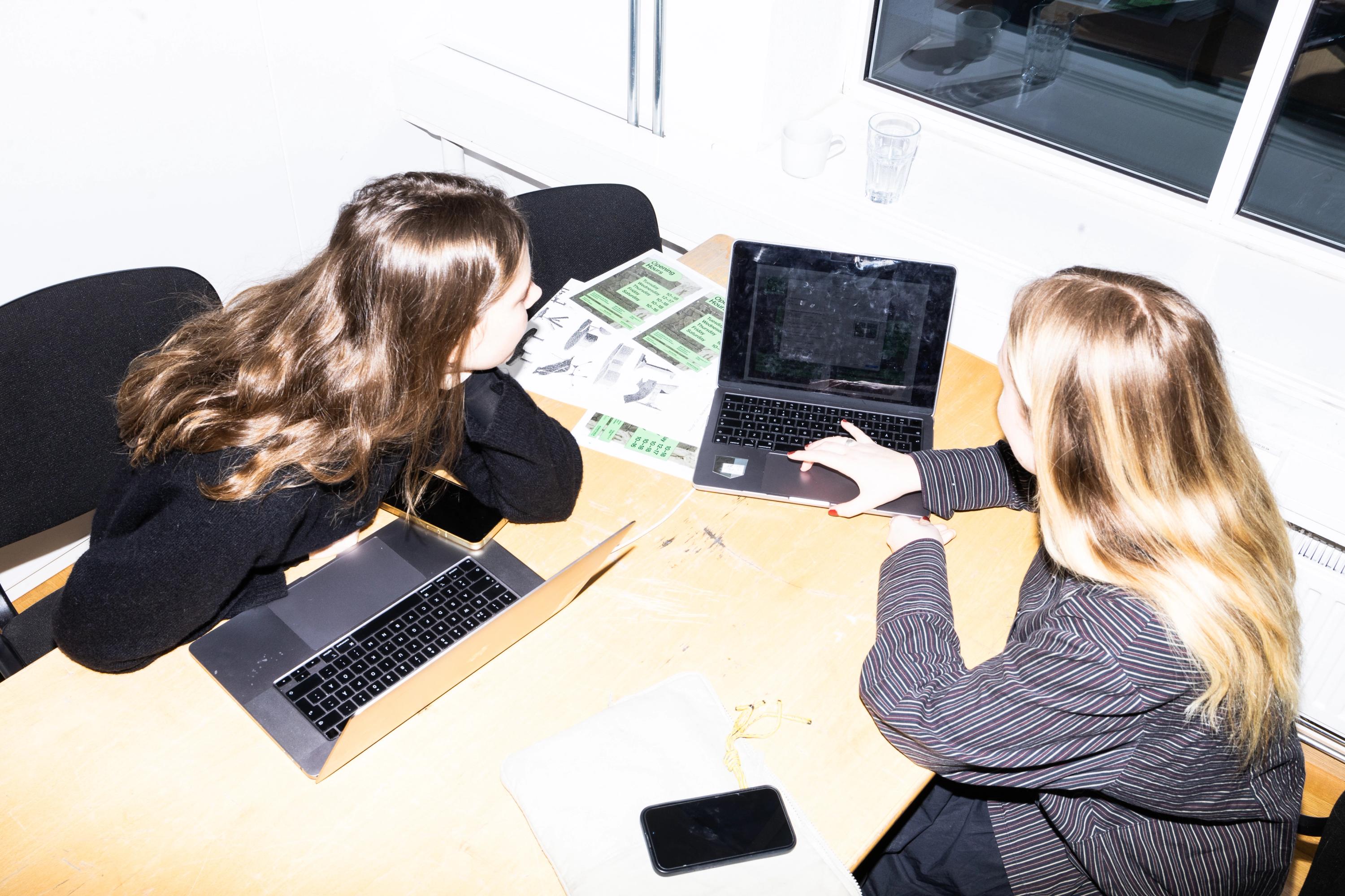 Två personer sitter vid ett bord med laptops och tittar tillsammans på en skärm, med utskrifter bredvid, fotograferat ovanifrån.