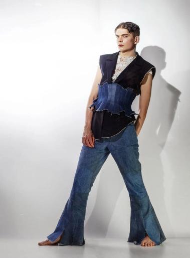 Barfota modell lutar sig mot en vägg i ärmlös mörk väst, spetstopp, jeanskorsett och utsvängda jeans.