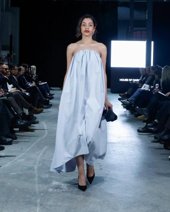 Modell på catwalk i axelbandslös ljusblå klänning med vid, asymmetrisk fåll och svarta klackar; publik på båda sidor.