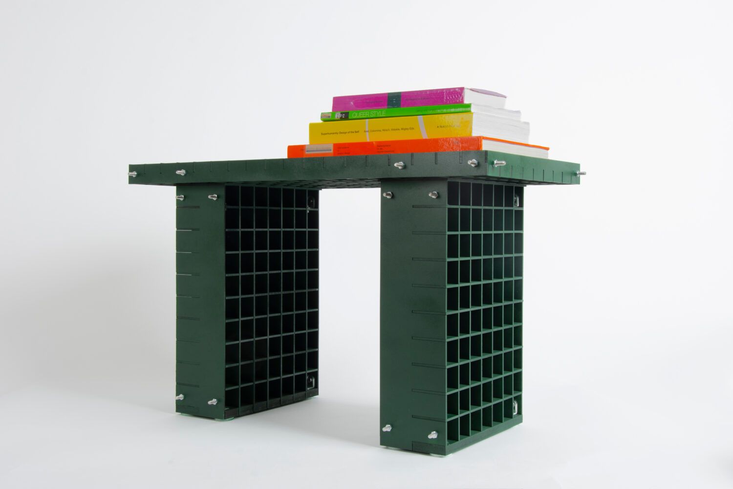Mörkgrön sideboard av hopskruvade, rutade plastpaneler, med en trave färgglada böcker ovanpå, mot vit bakgrund.