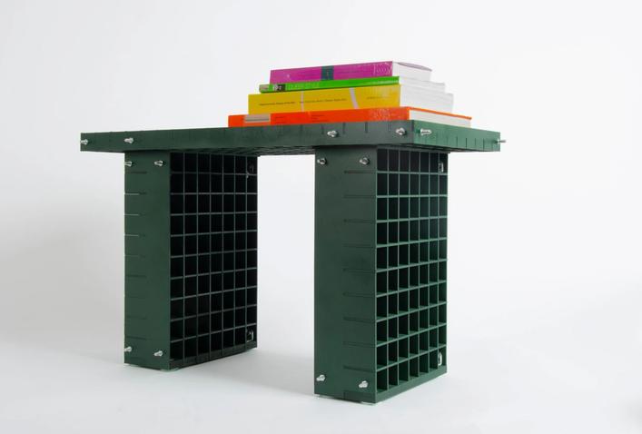 Mörkgrön sideboard av hopskruvade, rutade plastpaneler, med en trave färgglada böcker ovanpå, mot vit bakgrund.