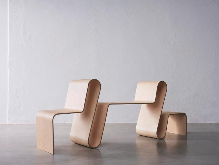 Bänk av böjd ljus plywood i ett stycke, med sitsar i olika höjder som bildar en sammanhängande form, på betonggolv.