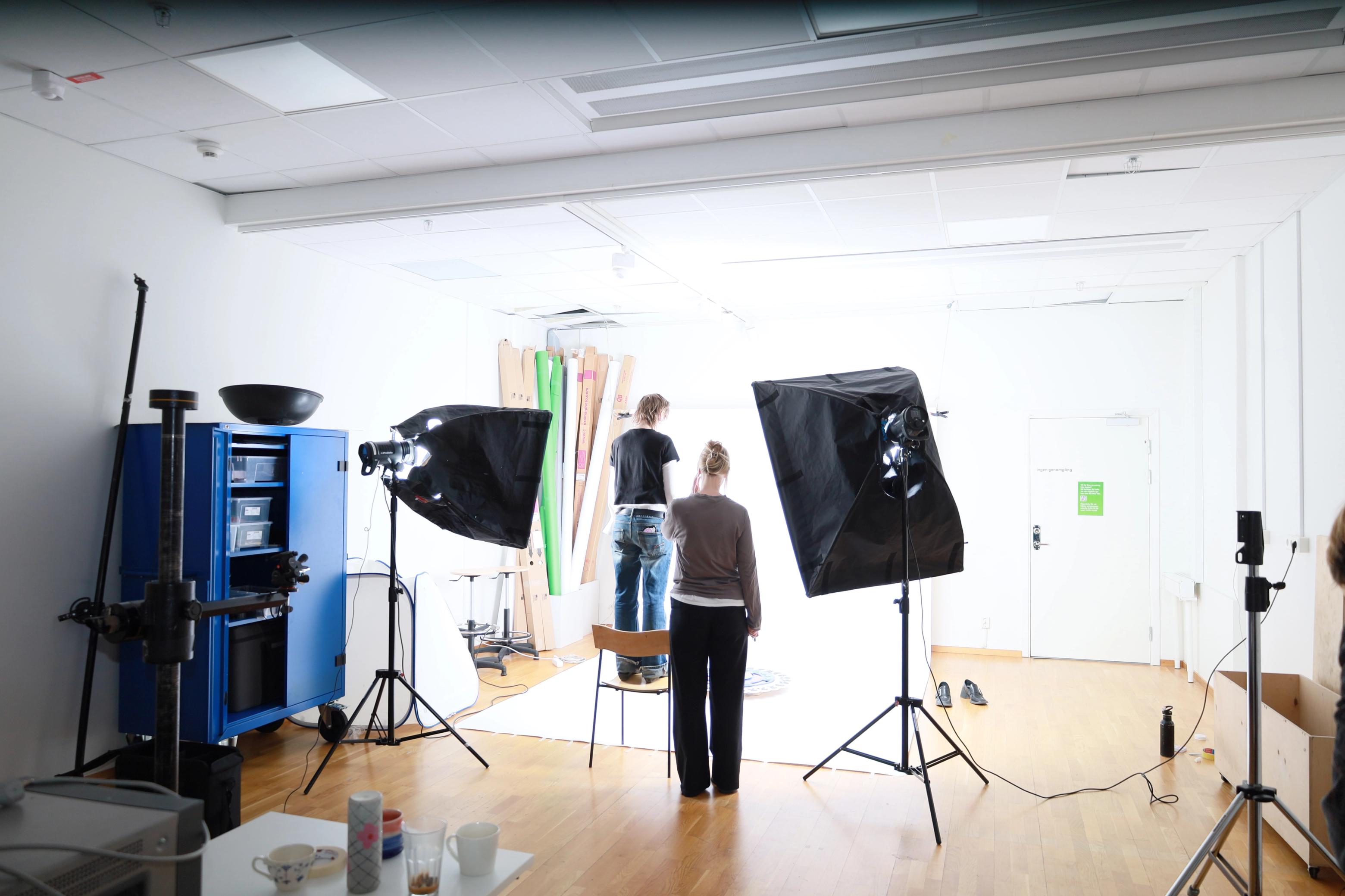 Två personer förbereder en fotografering i en ljus fotostudio med softboxar, stativ och bakgrundsrullar.