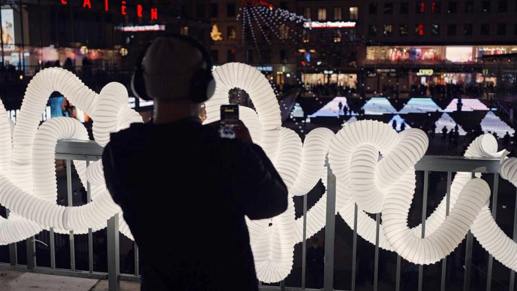 Person med hörlurar fotograferar en upplyst installation av vita slingrande rör längs ett räcke, över ett torg på kvällen.