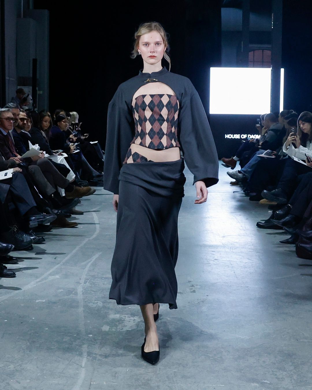 Modell på catwalk i mörkblå topp med vida ärmar och utskärning som visar ett rombmönster, lång kjol och svarta klackar; publik på båda sidor.