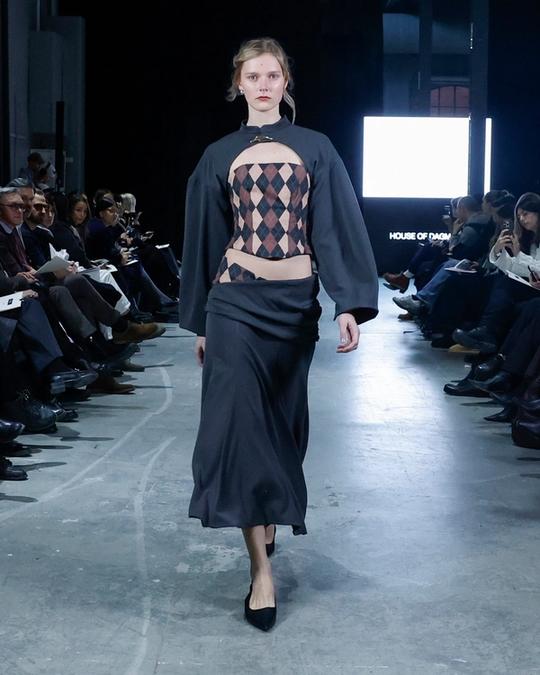Modell på catwalk i mörkblå topp med vida ärmar och utskärning som visar ett rombmönster, lång kjol och svarta klackar; publik på båda sidor.