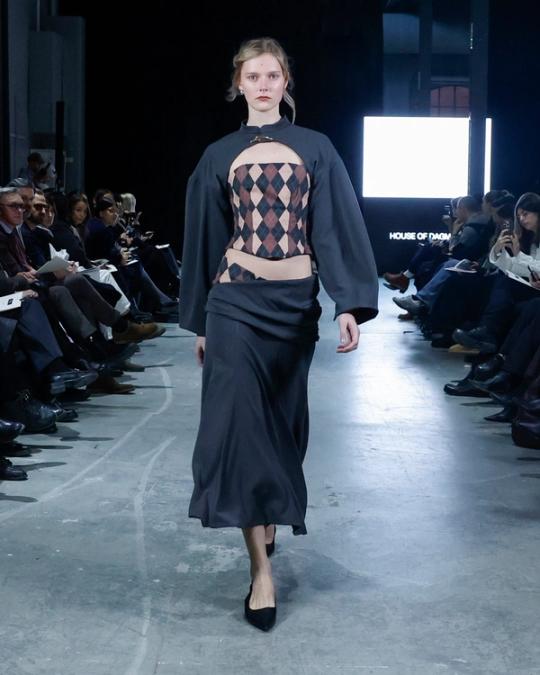 Modell på catwalk i mörkblå topp med vida ärmar och utskärning som visar ett rombmönster, lång kjol och svarta klackar; publik på båda sidor.