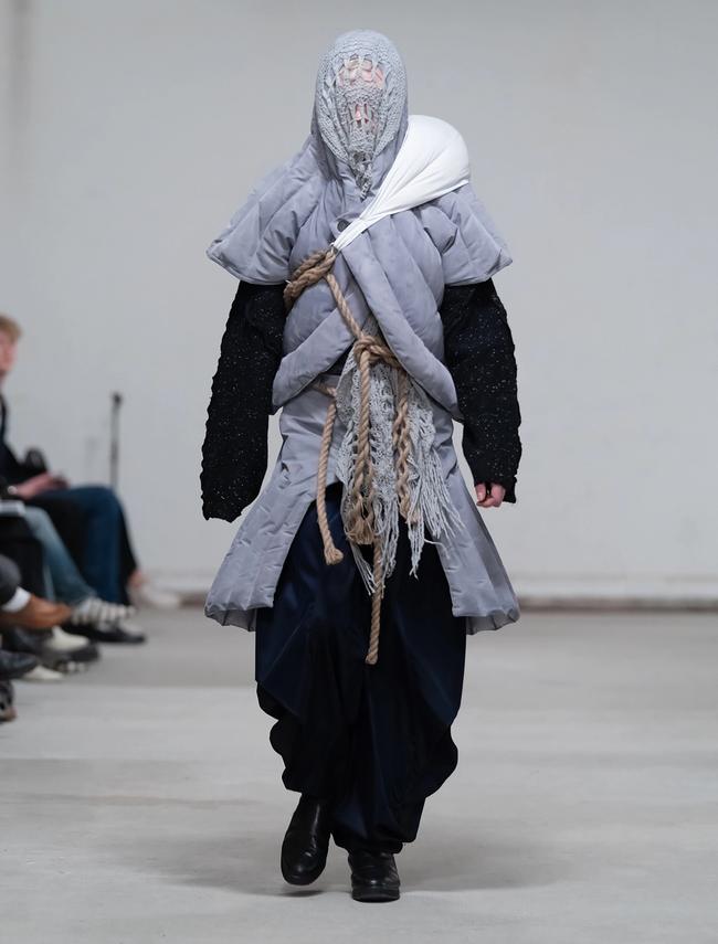 Modell på catwalk med ansiktet täckt av en grå stickad huva, i vadderad grå jacka lindad med rep och vida mörka byxor.