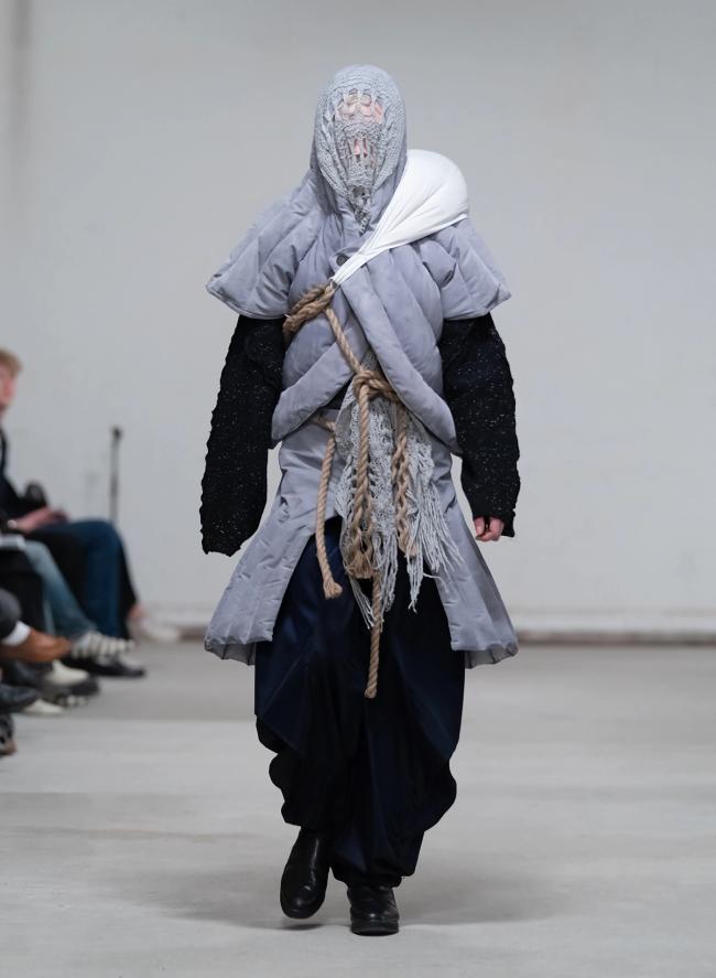 Modell på catwalk med ansiktet täckt av en grå stickad huva, i vadderad grå jacka lindad med rep och vida mörka byxor.