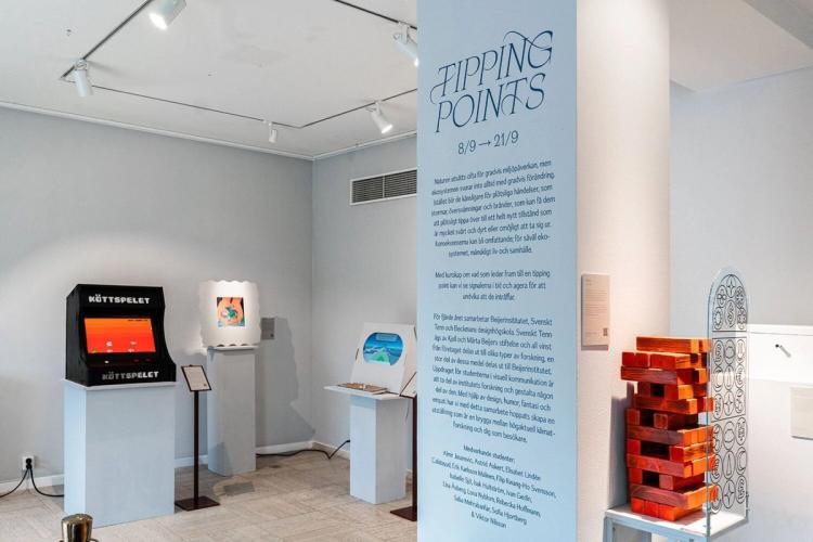 Utställningen "Tipping Points" med verk på vita podier, bland annat ett arkadspel och ett torn av träklossar, och en textvägg.