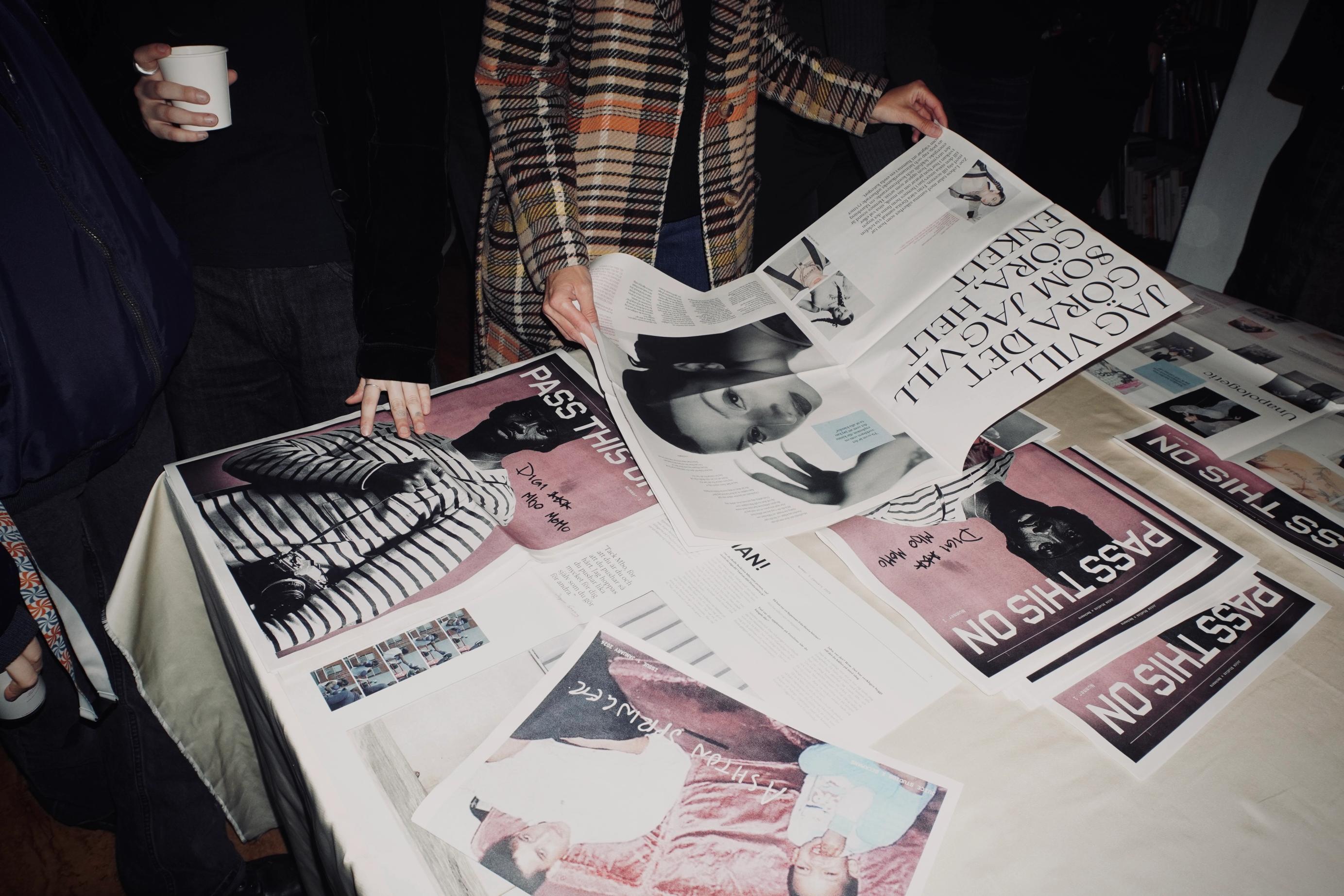 Personer bläddrar i tidningar gjorda som studentprojekt, utlagda på ett bord; en i rutig kappa slår upp ett nummer.