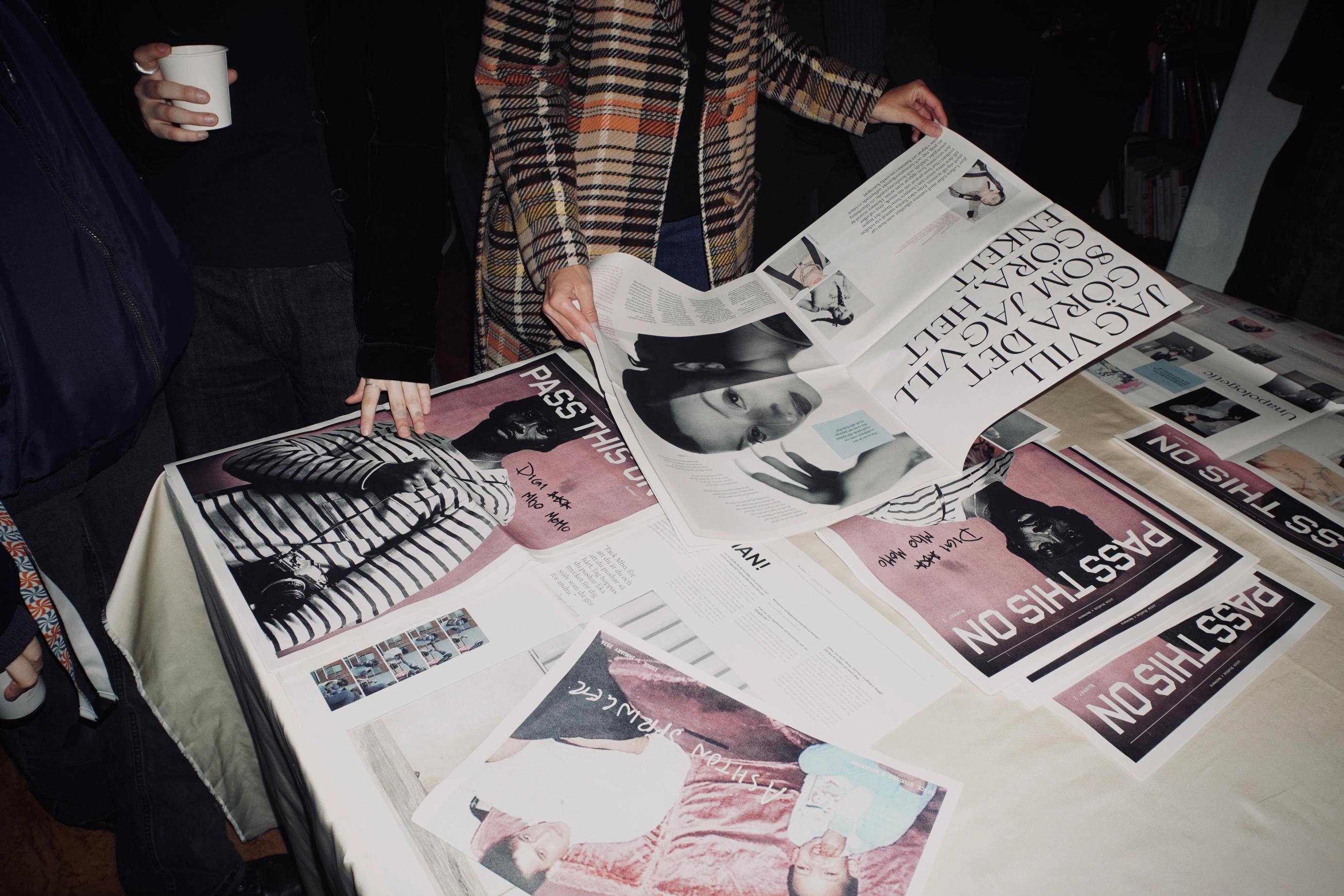Personer bläddrar i tidningar gjorda som studentprojekt, utlagda på ett bord; en i rutig kappa slår upp ett nummer.