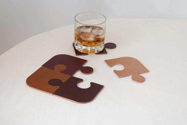 Glasunderlägg formade som pusselbitar i brunt läder, några ihoplagda, ett under ett whiskyglas med is.