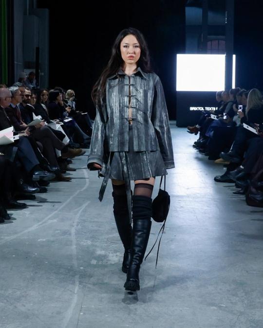 Modell på catwalk i grå metallisk jacka, matchande minikjol och svarta knähöga stövlar; publik sitter på båda sidor.