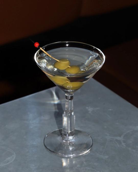Martiniglas med gröna oliver på en cocktailpinne, på en grå bänkskiva.