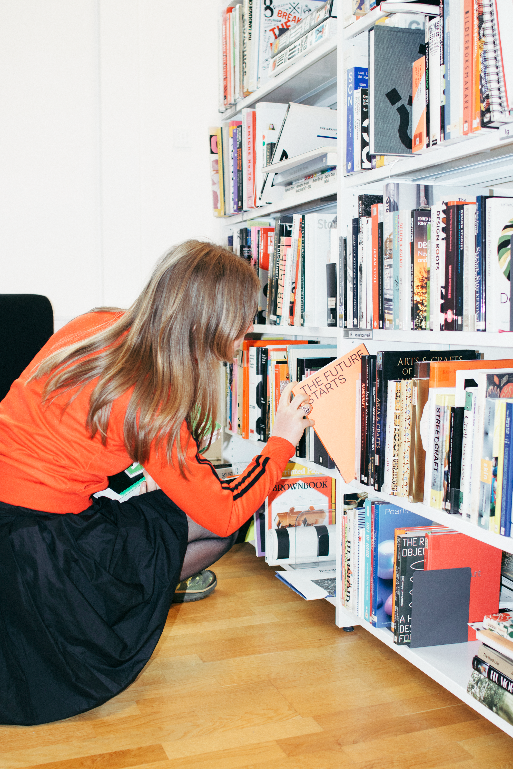 Person i orange jacka sitter på huk och tar fram boken "The Future Starts" ur en fullsatt bokhylla.