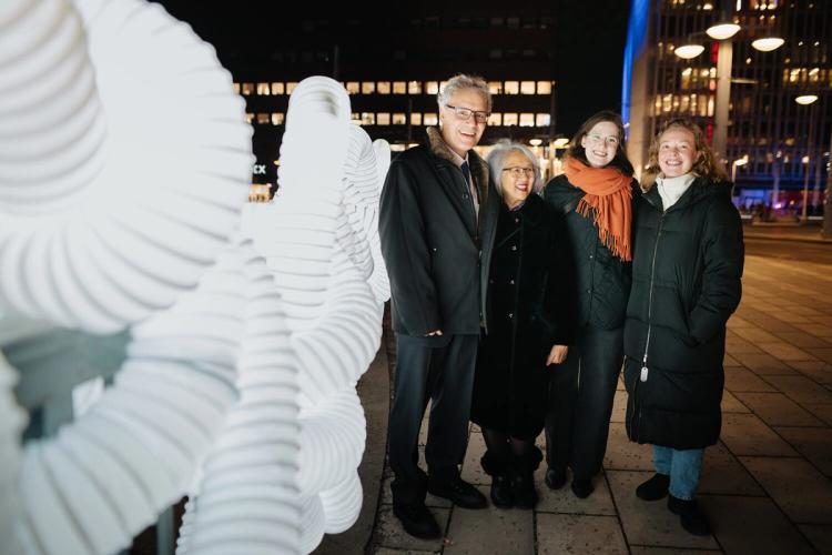 Fyra leende personer i vinterrockar står bredvid en upplyst installation av vita slingrande rör, i staden på kvällen.
