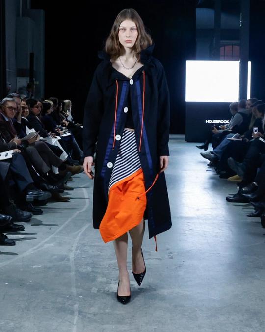 Modell på catwalk i lång mörk kappa över randigt och orange lager med snören, och svarta klackar; publik på båda sidor.