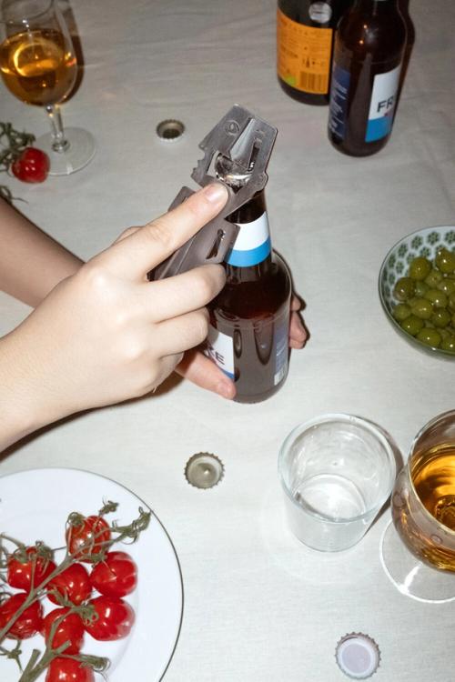 Händer öppnar en ölflaska med en metallkapsylöppnare formad som en liten figur, på ett bord med glas, oliver och körsbärstomater.