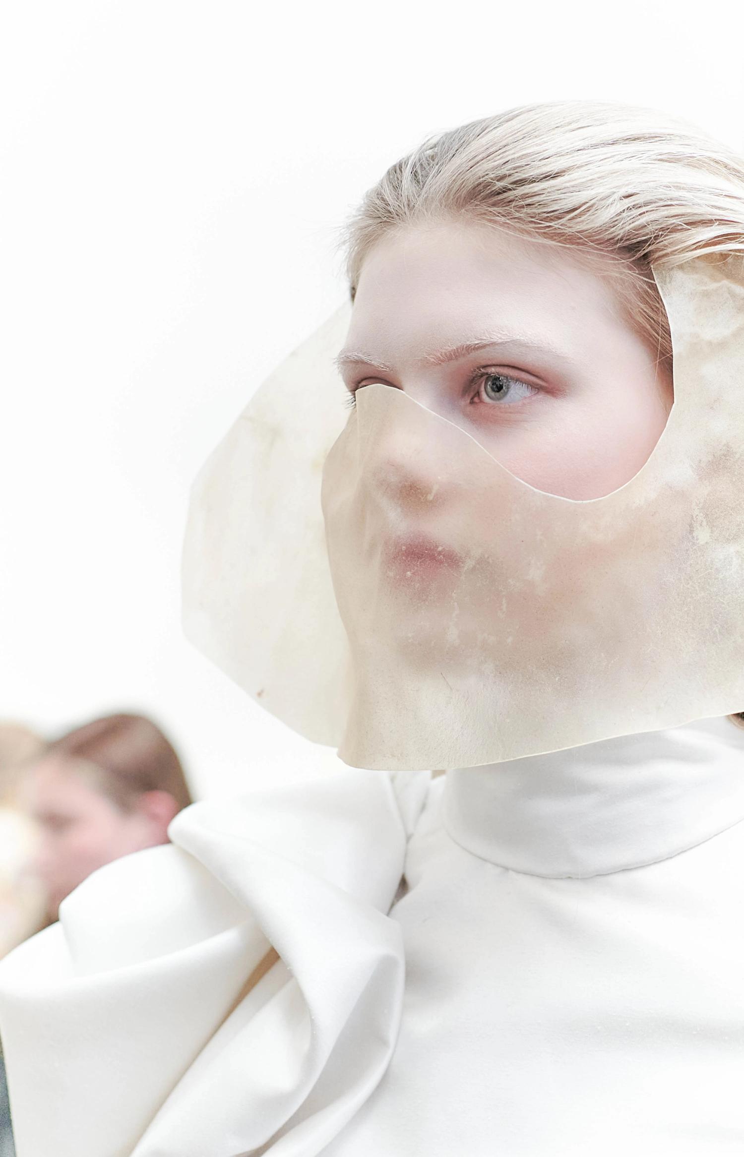 Modell i vitt plagg med en halvgenomskinlig mask som täcker nedre delen av ansiktet, med en öppning för ögonen.
