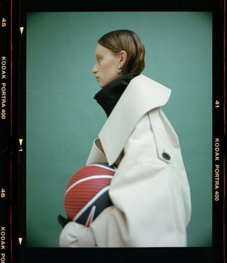 Profilporträtt på film av en modell i stor beige trenchcoat som håller en röd boll under armen, mot grön bakgrund.