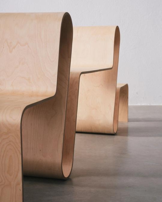 Närbild på stolar av böjd ljus plywood i ett stycke, uppställda på rad på ett betonggolv.