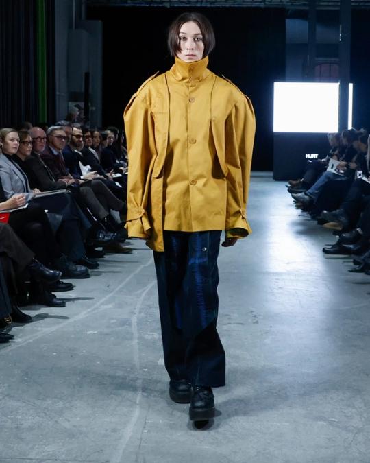 Modell på catwalk i stor senapsgul jacka med hög krage, vida mörkblå byxor och svarta skor; publik på båda sidor.