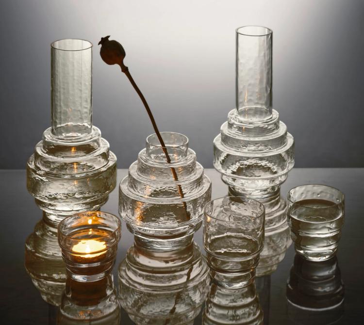 Hamrade glasvaser, glas och ett värmeljus i staplade former, en vas med en torkad vallmokapsel, på en blank yta.