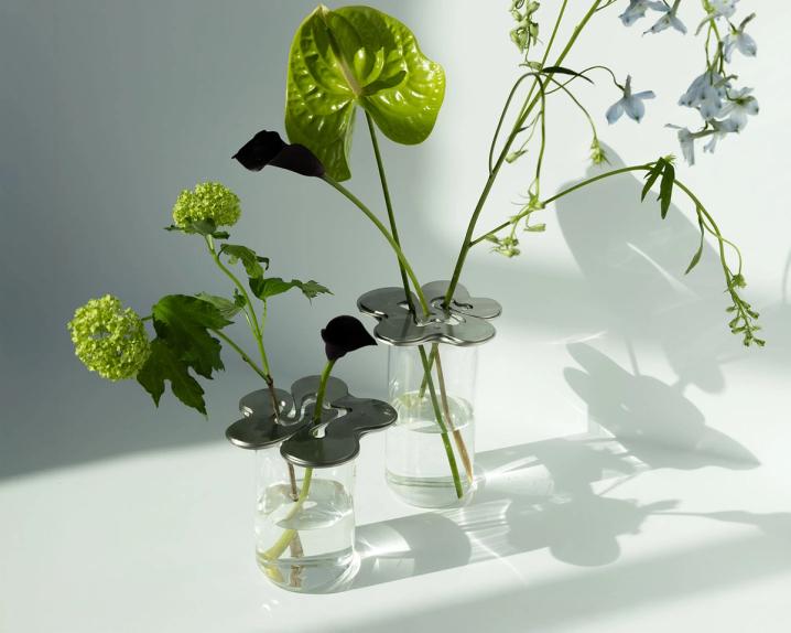 Två glasvaser med blomformade metallock som håller gröna och mörklila blommor och stjälkar, i solljus som kastar skuggor.