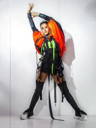 Modell poserar med armarna över huvudet i orange puffjacka, svart sele med neongröna detaljer, höga snörstövlar och sneakers.