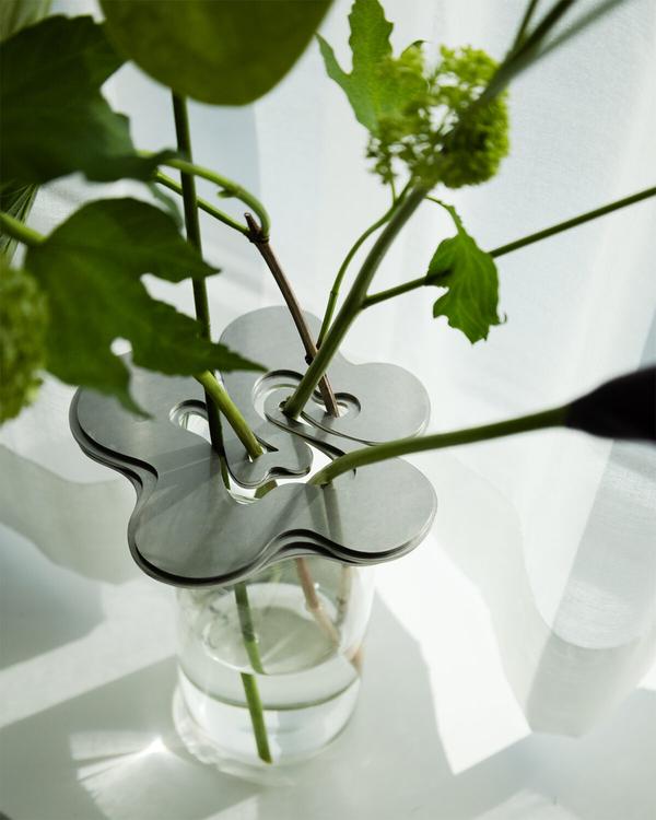 Närbild på en glasvas med blomformat metallock som håller gröna stjälkar och blad, framför en vit gardin.