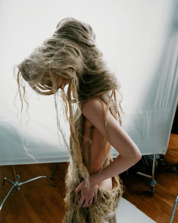Modell i fotostudio med huvud och kropp täckta av ett plagg av långa, hårliknande fibrer, mot vit bakgrundsduk.