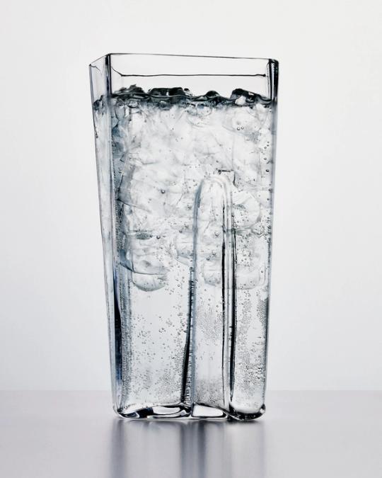 Högt fyrkantigt glas fyllt med kolsyrat vatten och isbitar, mot ljusgrå bakgrund.
