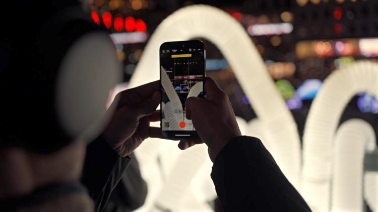 Person med hörlurar filmar en upplyst installation av vita slingrande rör med sin mobil, stadsljus i bakgrunden.
