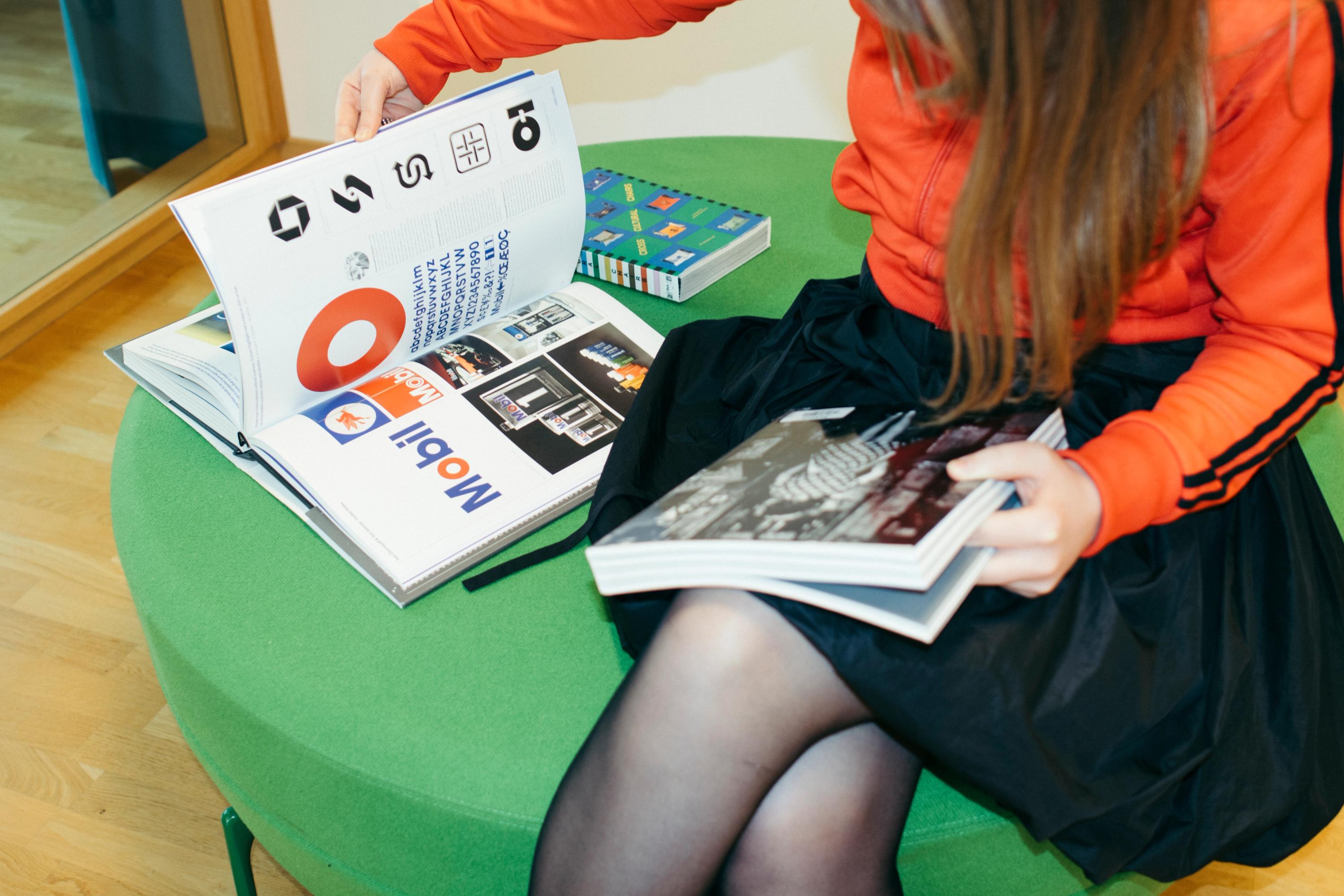 Person i orange jacka sitter på en grön pall och bläddrar i böcker om grafisk design och logotyper.